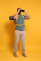 Happy tourist with backpack looking through binoculars on yellow background