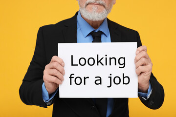 Unemployed senior man holding cardboard sign with phrase Looking For A Job on yellow background, closeup