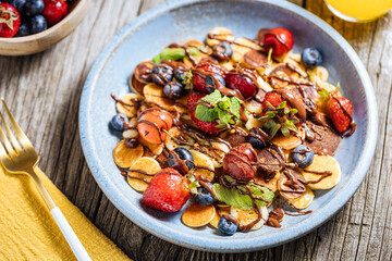 Small American pancakes mixed with seasonal fruit like strawberries, blueberries and cherries. Dipped with melted chocolate, placed on a rustic wooden board. 