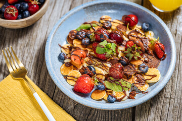 Small American pancakes mixed with seasonal fruit like strawberries, blueberries and cherries. Dipped with melted chocolate, placed on a rustic wooden board. 
