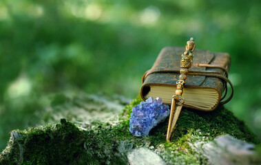 Ritual knife, amethyst druse, magic spell book on mossy stump, blurred natural green background. Phurba is three-sided knife, traditionally of Indo-Tibetan Buddhism for esoteric spiritual practice