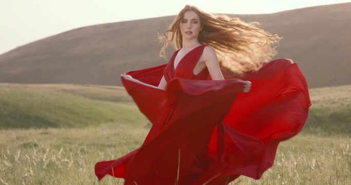 Slow motion, Caucasian woman in red dress in field spinning around her axis, developing hem of dress in wind. Hem of red dress opens smoothly in wind, like butterfly wings. Woman spinning in red dress