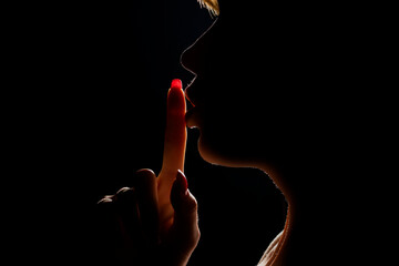 Silhouette of unknown woman with the face in the shadow holding finger on her lips on a black...