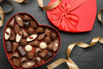 Heart shaped box with delicious chocolate candies and ribbon on black table, flat lay
