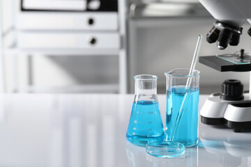 Different laboratory glassware with light blue liquid near microscope on table, space for text