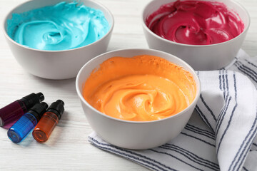 Bowls of different cream with food coloring and bottles of bright liquid on white wooden table