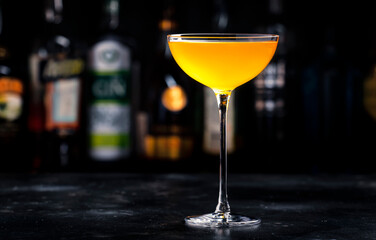 Golden alcoholic cocktail drink with whiskey, apple liqueur, ginger syrup, lemon juice, green basil and ice, dark bar counter background