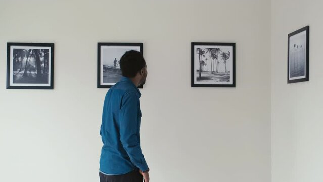 Young black man walking in art gallery and watching modern photos on walls while visiting exhibition