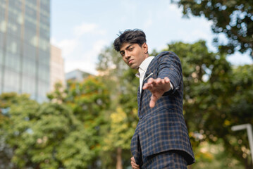 A stylish guy in a blue checkered suit in a serious face with a reaching his hand down gesture while standing outside, in the city. Sky, building and trees are shown in the background.