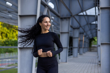 Young beautiful hispanic woman with curly hair jogging near stadium, woman in tracksuit smiling...