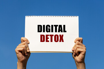 Digital detox text on notebook paper held by 2 hands with isolated blue sky background. This message can be used as business concept about digital detox.