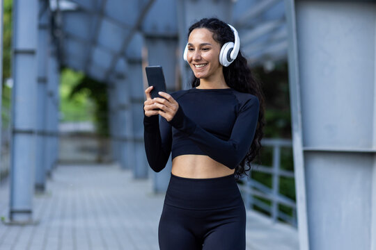 Hispanic sportswoman with headphones listening to online music and audio podcasts with books while jogging and doing active exercises, woman using app on phone, in tracksuit.