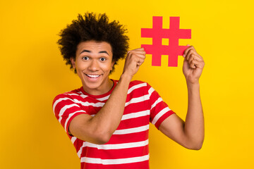 Photo of cheerful positive guy dressed red t-shirt smiling rising hash tag card isolated yellow color background