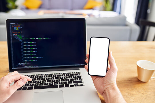 Man Working On Laptop At Home Holding Phone With Blank Screen