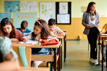 Obraz na płótnie Canvas Hispanic schoolgirl and her classmates writing in classroom.