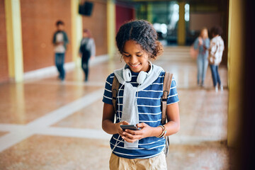 Happy black teenage girl using mobile phone in high school hallway.
