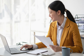 Asian businesswoman worker or manager in a yellow suit in a good mood using a laptop and calculator to calculate company income and expenses balance inside the office.