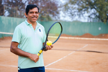 Happy indian senior man with racquet and ball by looking at camera at tennis court - concept of hobbies, successful and relaxation.
