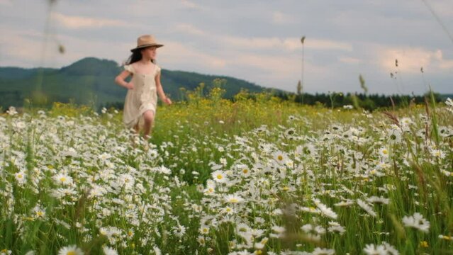 Selective focus of adorable smiling little girl kid in hat runs through field of daisies during sunset. Concept weekend in nature. Childhood and freedom, child in dress running through blooming meadow