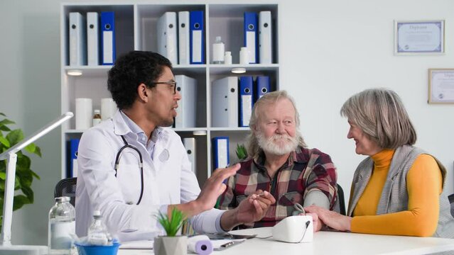 afro american male doctor measures pressure of an elderly male patient and consults old wife about state of health while sitting in a medical office