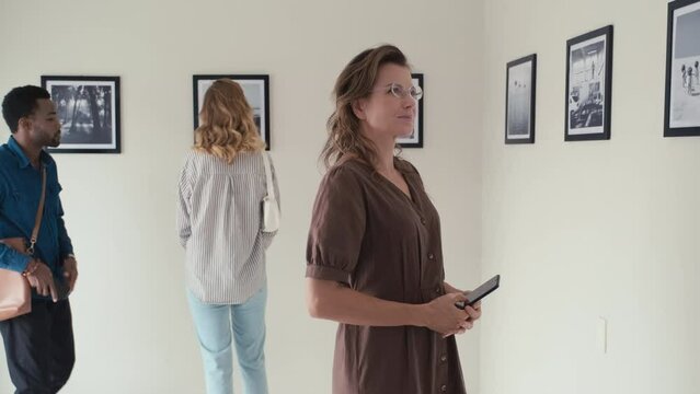 Medium long shot of woman watching photo exhibition in art gallery and taking pictures with mobile phone, other visitors walking behind