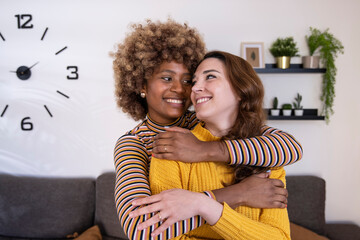 Beautiful and cheerful multiracial lesbian couple hugging and looking each other smiling. Two lovely and happy homosexual women embracing at home staring each other.