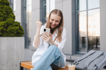 Successful woman using phone rejoices in winning success, looks at phone, dressed in casual clothes, sitting near office, businesswoman boss. Lucky girl look surprised, she win, triumph.