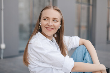 Happy blonde woman sitting on the bench near office building with wireless earbuds in ears and listening music, look at side. Girl look happy, she is smiling. Woman wear white shirt and blue jeans.