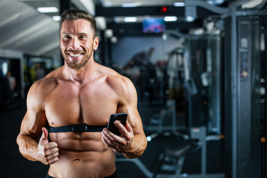 Strong, fit, athletic man in a gym with heart rate monitor around chest checking heart rate on cell phone with a big smile and thumb up. Good result, good training, happy face.