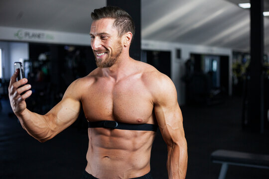 Strong, fit, athletic man in a gym with heart rate monitor around chest checking heart rate on cell phone with a big smile. Good result, good training