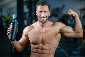 Fototapeta na wymiar Strong, fit, athletic, attractive man in a gym. Holding black shaker in one hand and shows muscles with other hand. Concept protein, water, health.