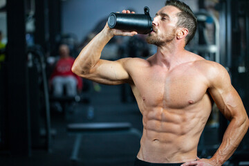 Strong, fit, athletic, attractive man in a gym. Drinking protein or water from the black shaker . - obrazy, fototapety, plakaty