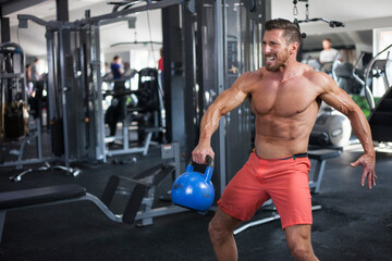 Strong muscular man swings blue kettlebell  with one hand in a gym. Powerful and strong face expression