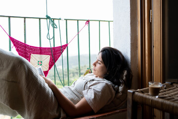 girl on balcony reading relaxed, woman in her twenties in rural house relaxing with a book, coffee with milk on a stool, brunette with skin and face dressed in a jumpsuit.