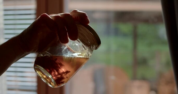 Moth larvae are soaked in liquid. A hand demonstrates a derivative of a tincture. Glare of light in a jar, cloudy liquid.