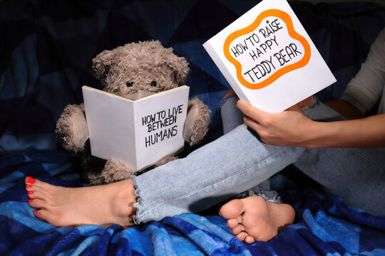 Girl in blue jeans sitting barefoot on a couch reading "How to raise a Happy Teddy bear" book, next to her sit Teddy Bear reading "How to live between Humans". Charming evening reading time concept.