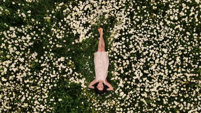 Aerial drone view of happy child girl enjoying beautiful fields daisies in summertime. Pretty relaxed little kid lying in flowers at sunny day. Carefree preteen is enjoying freedom on rural nature