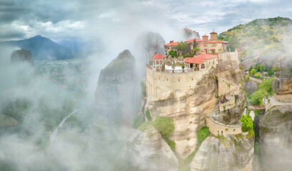 Meteora Varlaam Monastery Greece, rising out of the mist. Amazing mystical panoramic landscape. View of mountains and green forests against a dramatic sky with clouds. A UNESCO World Heritage Site.  