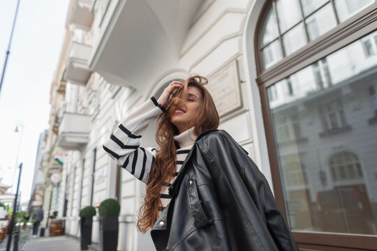 Happy beautiful young girl model brunette in fashionable clothes with a striped sweater and a leather black jacket walks on the street near vintage white buildings. Travel in Europe