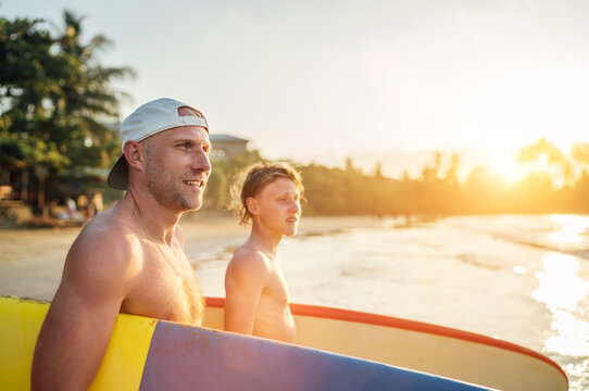Smiling man with Young teen boy son with surfboards go to the sea for surfing. They have a winter vacation and enjoying a beautiful sunset light. Family active vacation concept