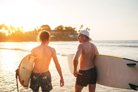 Father with teenager son walking with surfboards by sandy ocean beach with palm trees on background lightened with sunset sun They have conversation and enjoy summertime Family active vacation concept
