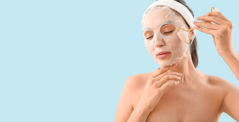 Young woman with applied sheet facial mask and massage roller on light blue background with space...