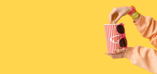 Female hands with stylish wristwatches, bucket of popcorn and 3D glasses on yellow background with...