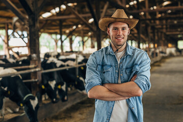Portrait of a happy farmer standing in a barn full of cows. - obrazy, fototapety, plakaty