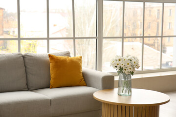 Interior of light living room with grey sofa and wooden coffee table near big window
