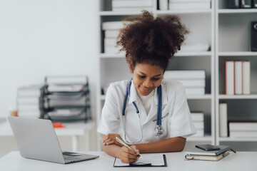 Young African american female doctor using smartphone and working with computer while sitting at the clinic room.