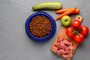 Bowl with dry pet food, raw meat and natural products on color wooden background