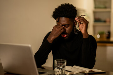 A tired african american freelancer is sitting at home at night and rubbing his eyes.