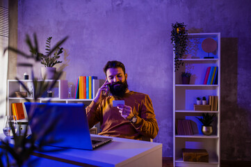 A happy entrepreneur is sitting in a neon blue and purple lighted home office with a credit card in his hands and talking on the phone.