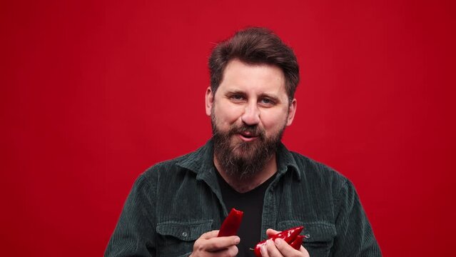 Bearded man eating red hot chilli pepper against red studio background. Spicy taste. Lover of hot, spicy food. Concept of food, vegetables, taste, grocery, human emotions. Ad